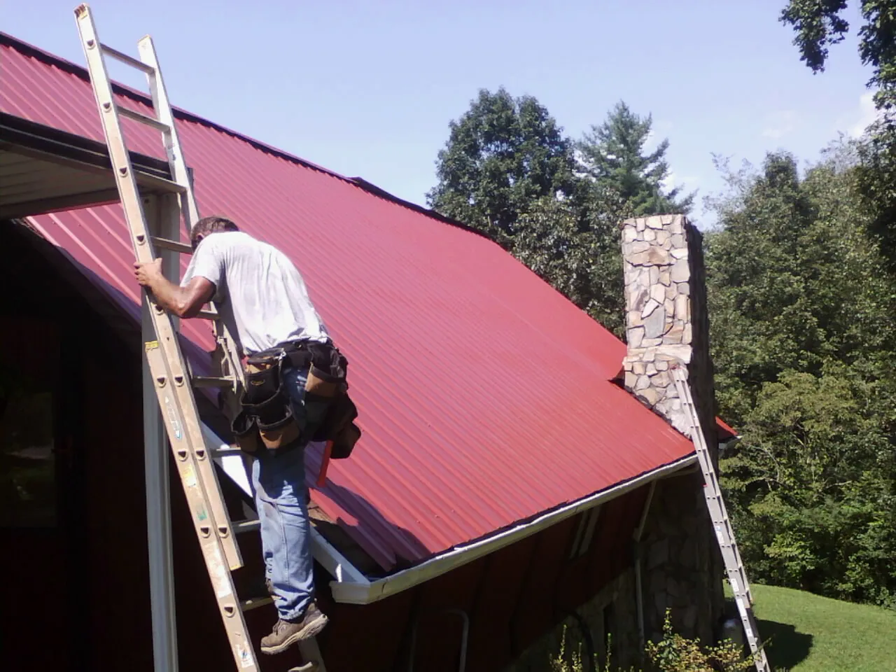 Roofer on a ladder beside a red metal roof and stone chimney — Soffit & Fascia Repair by Roofing Brothers in Plain City